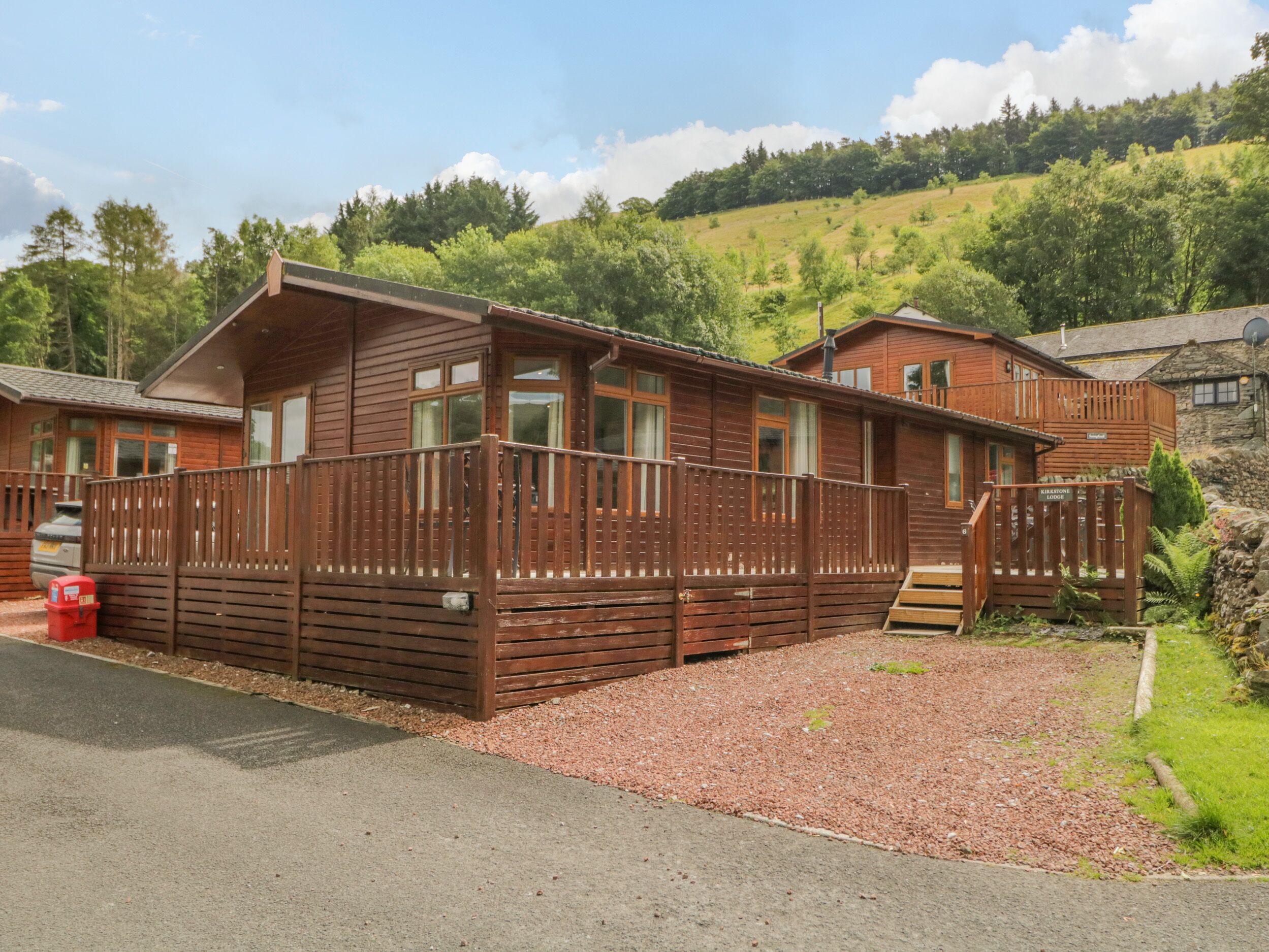 A wooden cabin with a balcony, a gravel driveway, and a stone wall.