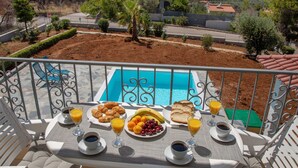 Un balcón con una mesa lista para el desayuno, con vista a una piscina, un edificio moderno y un paisaje montañoso.