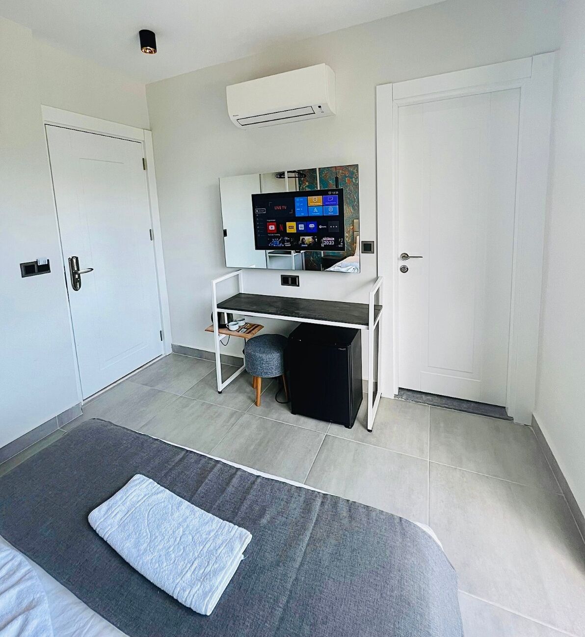 A modern hotel room with a bed, a desk with a computer, an air conditioner, and a TV.