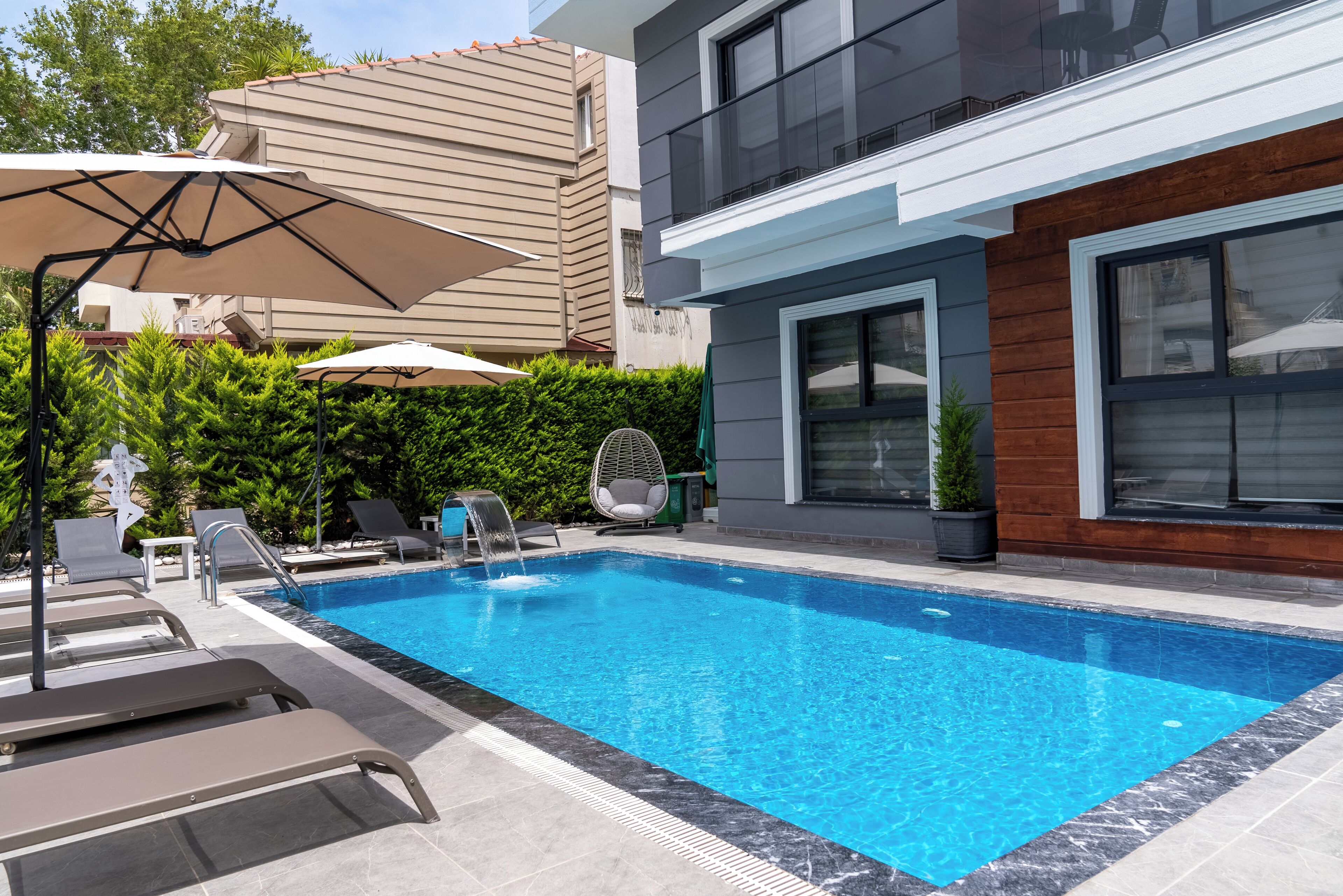 A swimming pool with lounge chairs and umbrellas, surrounded by a hedge and modern buildings.