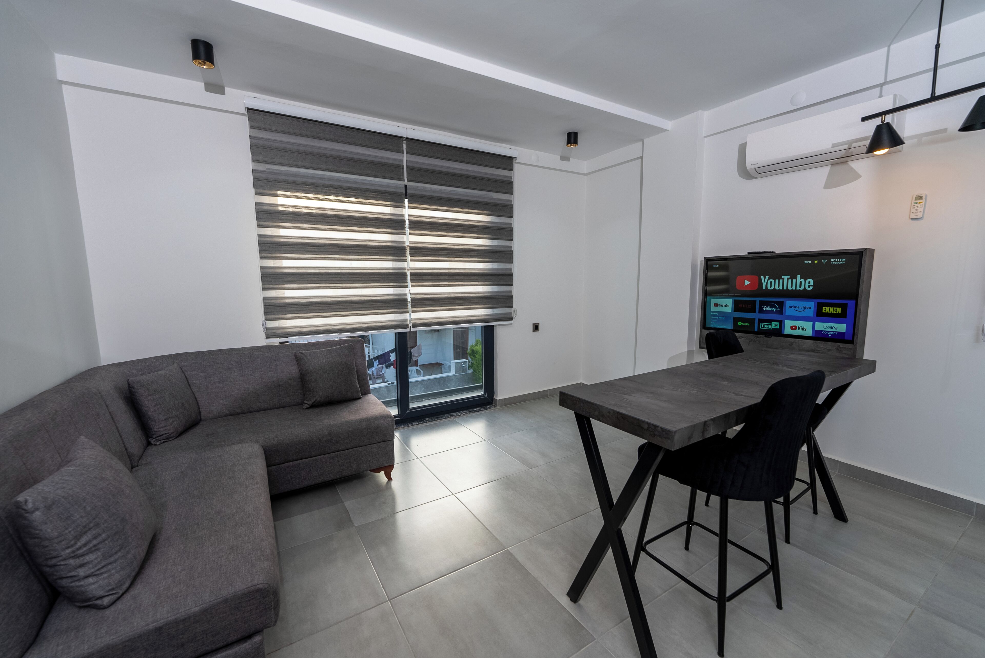 A modern living room with a grey sectional sofa, a wooden dining table with black chairs, a flat-screen TV on the wall, and a large window with blinds.