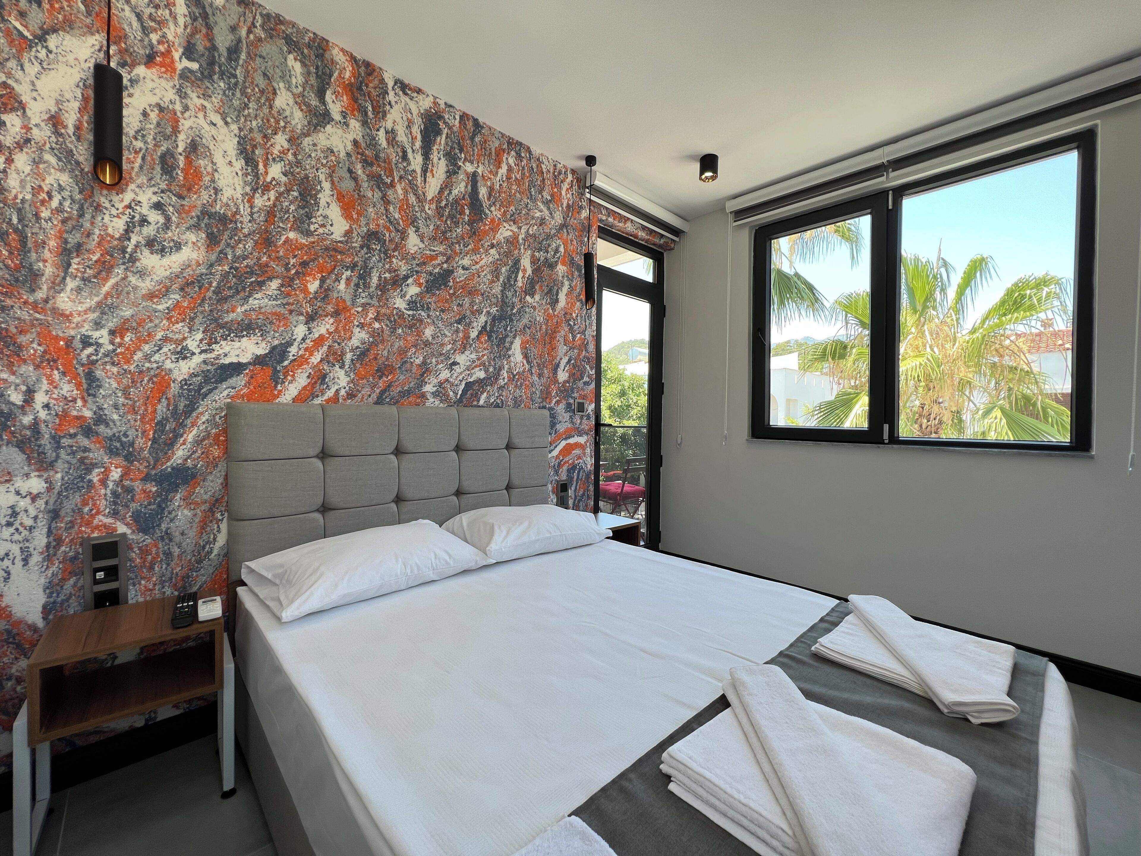 Una habitación de hotel moderna con una cama grande, un muro con diseño de mármol, un ventanal con vistas a palmeras y una mesita de noche con lámpara.