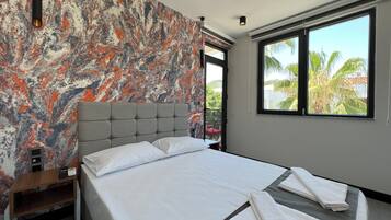 Una habitación de hotel moderna con una cama grande, un muro con diseño de mármol, un ventanal con vistas a palmeras y una mesita de noche con lámpara.