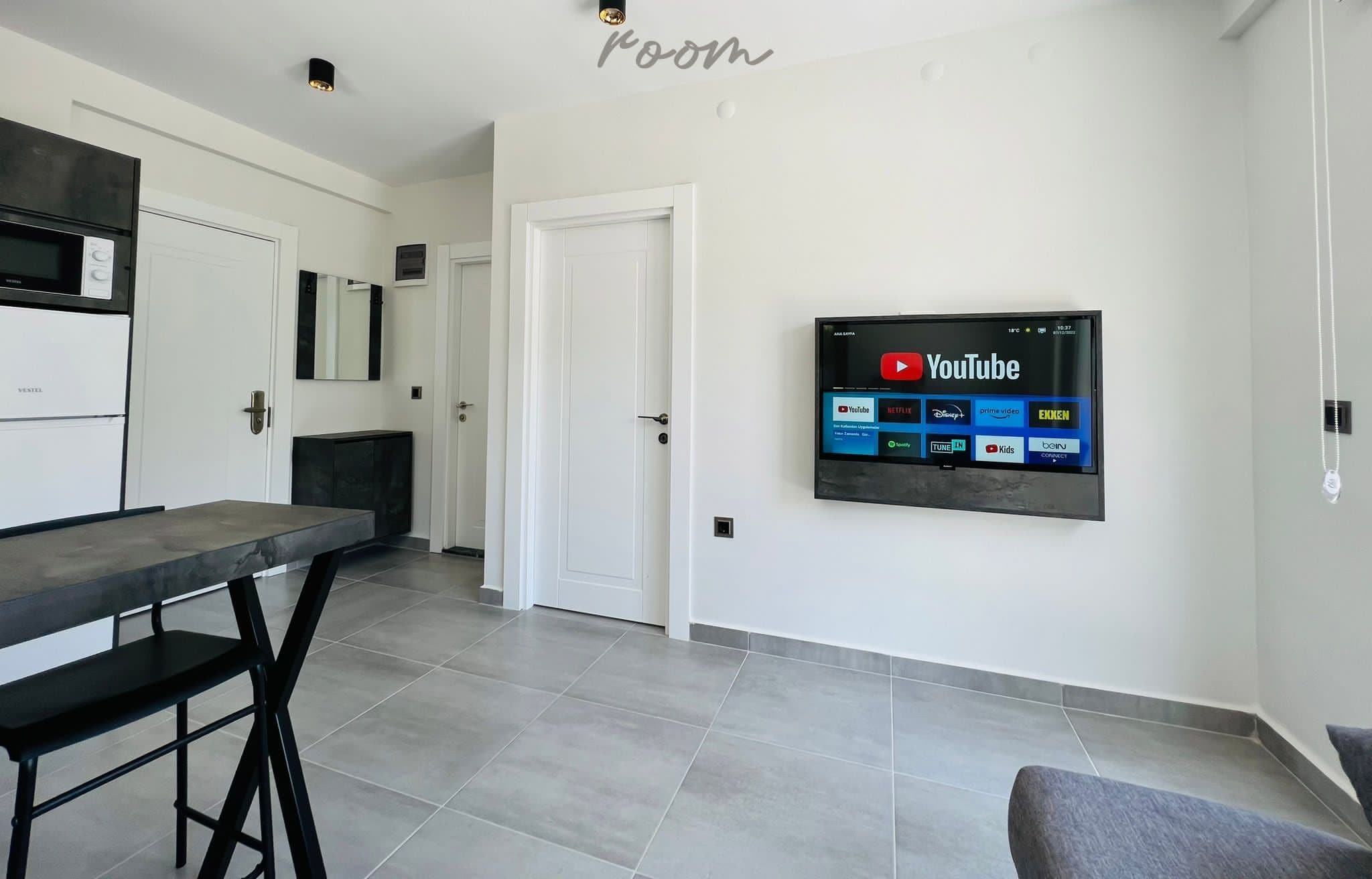 A modern living room with a flat-screen TV mounted on the wall displaying YouTube channels, a black dining table with a bench, and a white door leading to another room.