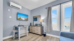 A modern bedroom with a desk, a dresser, a large window with curtains, and a beach-themed painting.