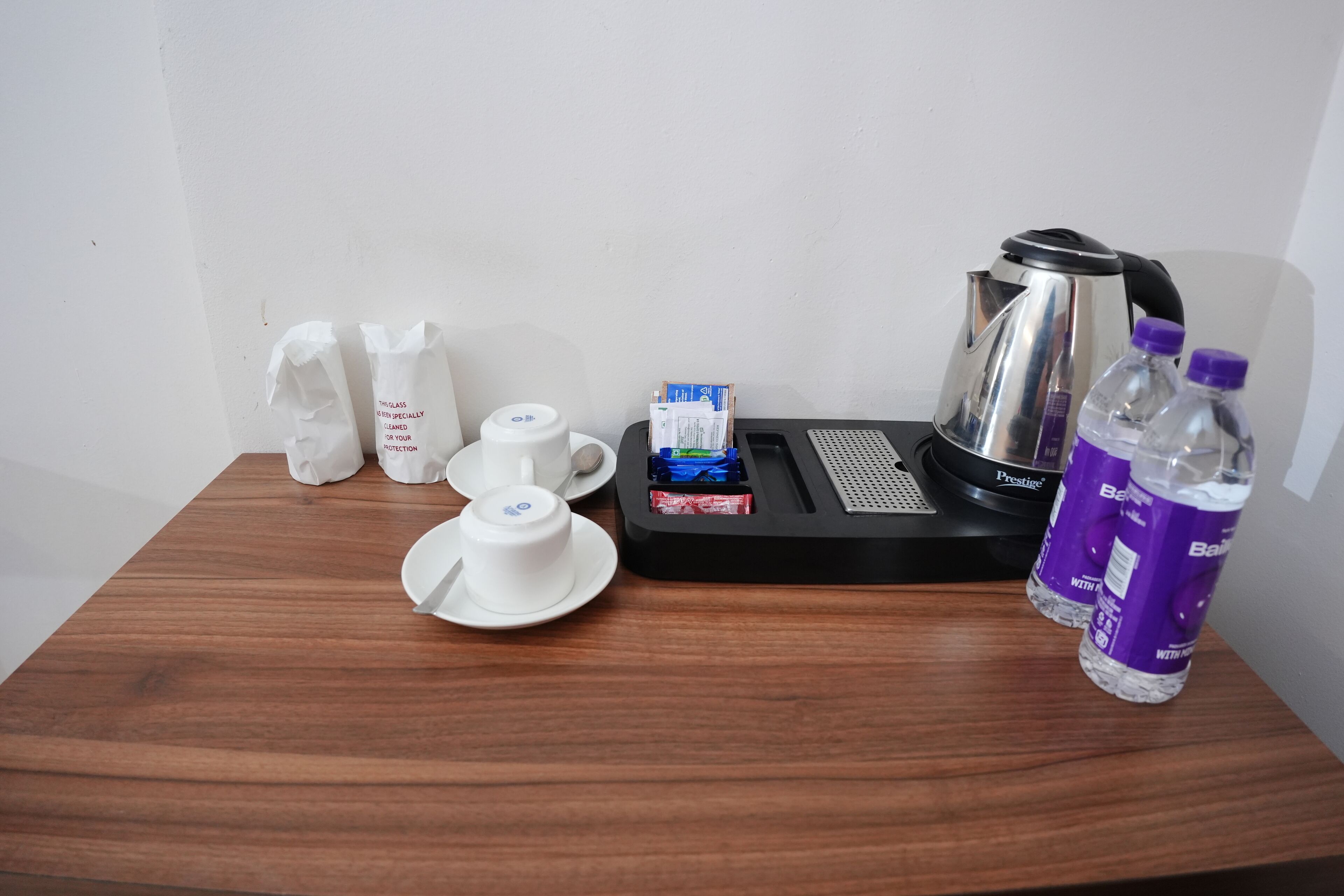 A wooden table with a coffee maker, two cups, a box of tissues, a tissue box, and two bottles of water.