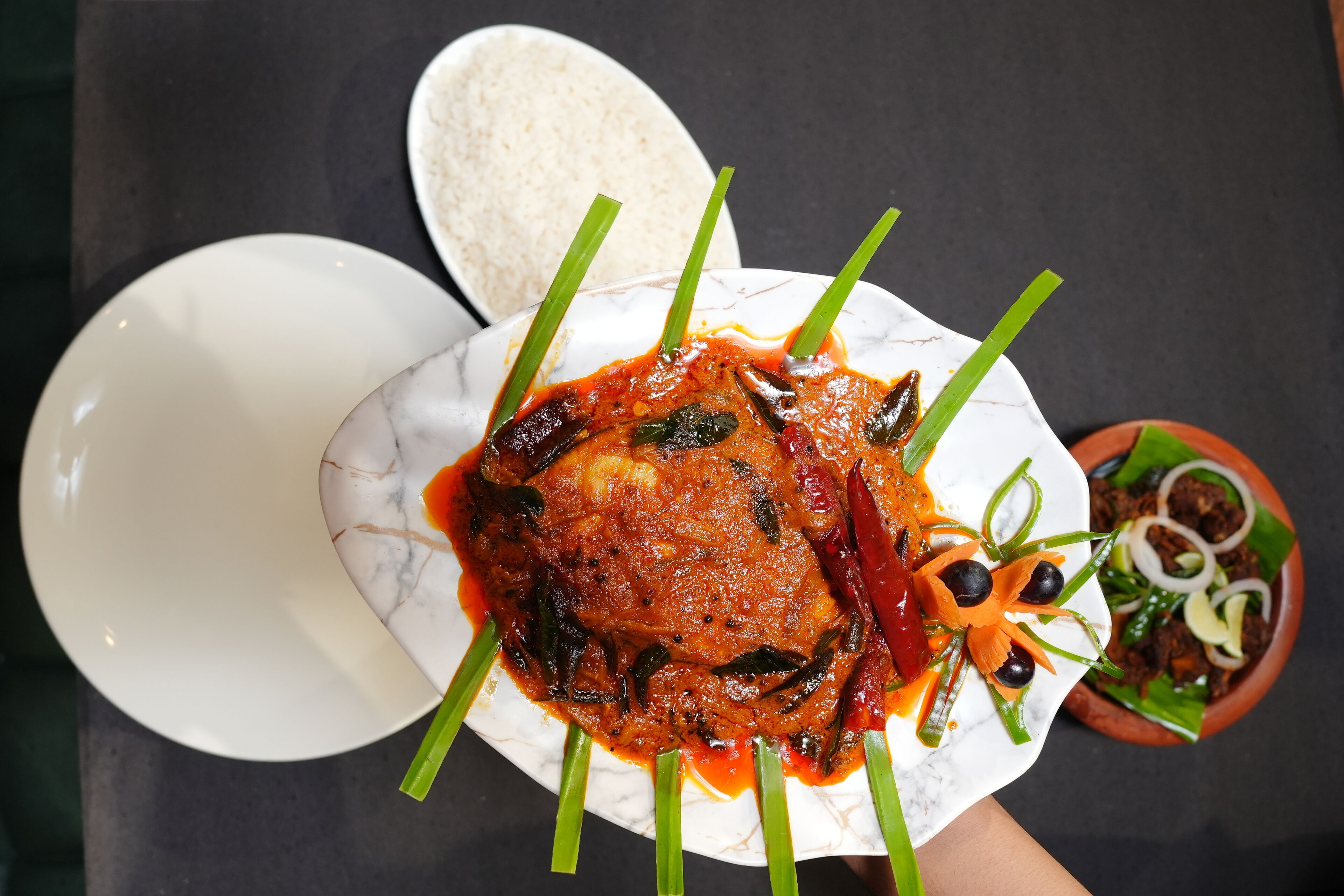 A plate of fish in a red sauce garnished with green onions and chili peppers, accompanied by a bowl of rice and a side dish of sliced onions and carrots.