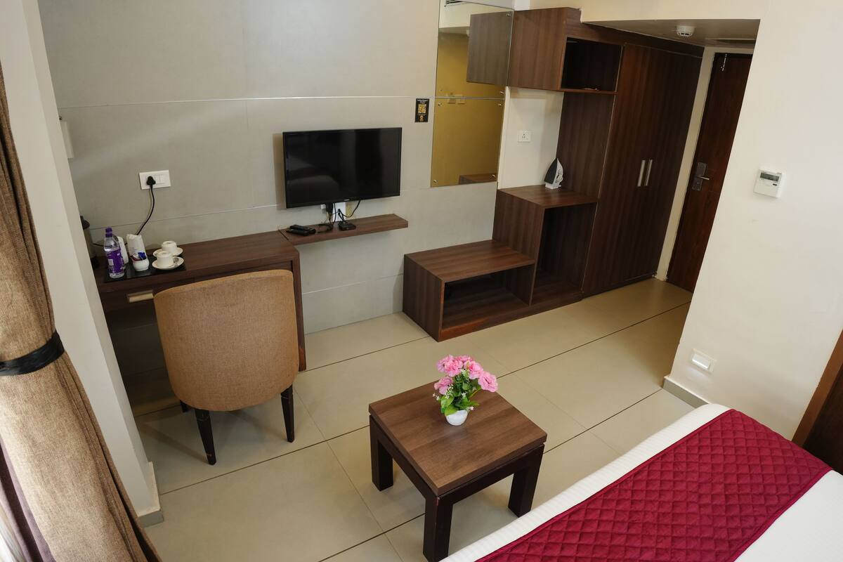 A modern living room with a flat-screen TV, wooden furniture, a small table with a vase of flowers, and a red bedspread.