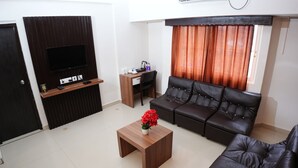 A living room with a black leather sofa, a wooden coffee table with a vase of red flowers, a flat-screen TV mounted on a wooden panel, a small wooden desk with a chair, and a window with orange curtains. - Hotel Palmyra Grand Inn (Kanayannur)