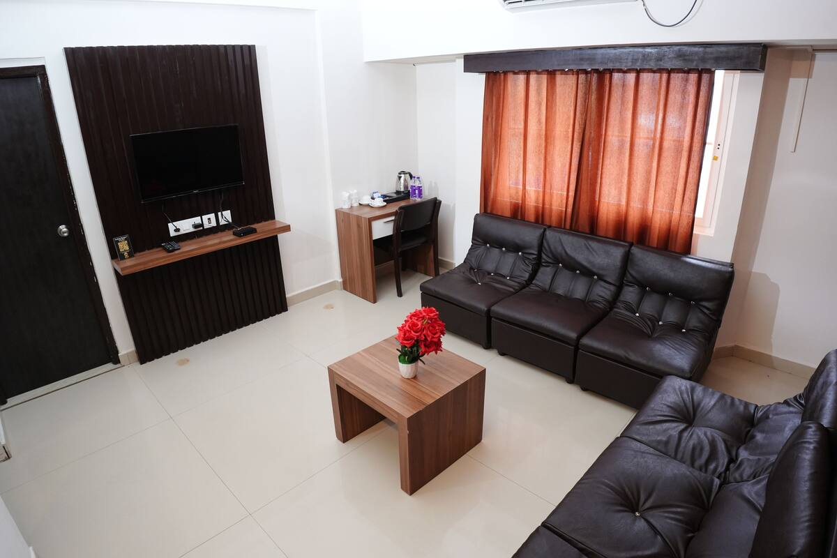 A living room with a black leather sofa, a wooden coffee table with a vase of red flowers, a flat-screen TV mounted on a wooden panel, a small wooden desk with a chair, and a window with orange curtains.