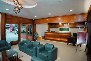 Zona de recepción de hotel con un mostrador de madera, un reloj digital y una barra de recepción. Hay varias sillas tapizadas en verde y una mesa de centro de vidrio en el área de estar.