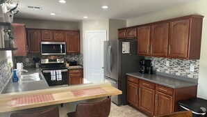 A kitchen with wooden cabinets, stainless steel appliances, a tiled backsplash, and a bar area with stools. - AZ Sun & Serenity: 4BR home w/Heated Pool, BBQ Island, Prime location– Sleeps 12 (Surprise)