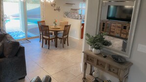 A living room with a sofa, a coffee table, and a dining area with chairs and a table. There is a television mounted on the wall and a decorative mirror above it. - AZ Sun & Serenity: 4BR home w/Heated Pool, BBQ Island, Prime location– Sleeps 12 (Surprise)