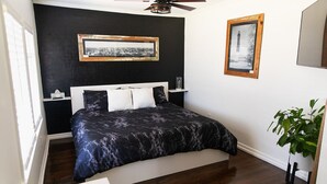 A bedroom with a bed, a ceiling fan, a nightstand, a framed picture, and a potted plant.