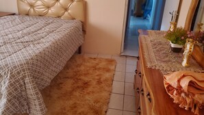 A bedroom with a tufted headboard, a patterned bedspread, a fluffy rug, a wooden dresser with a lace tablecloth, and a potted plant. - Mckay's Home Away from Home (Hatfield)