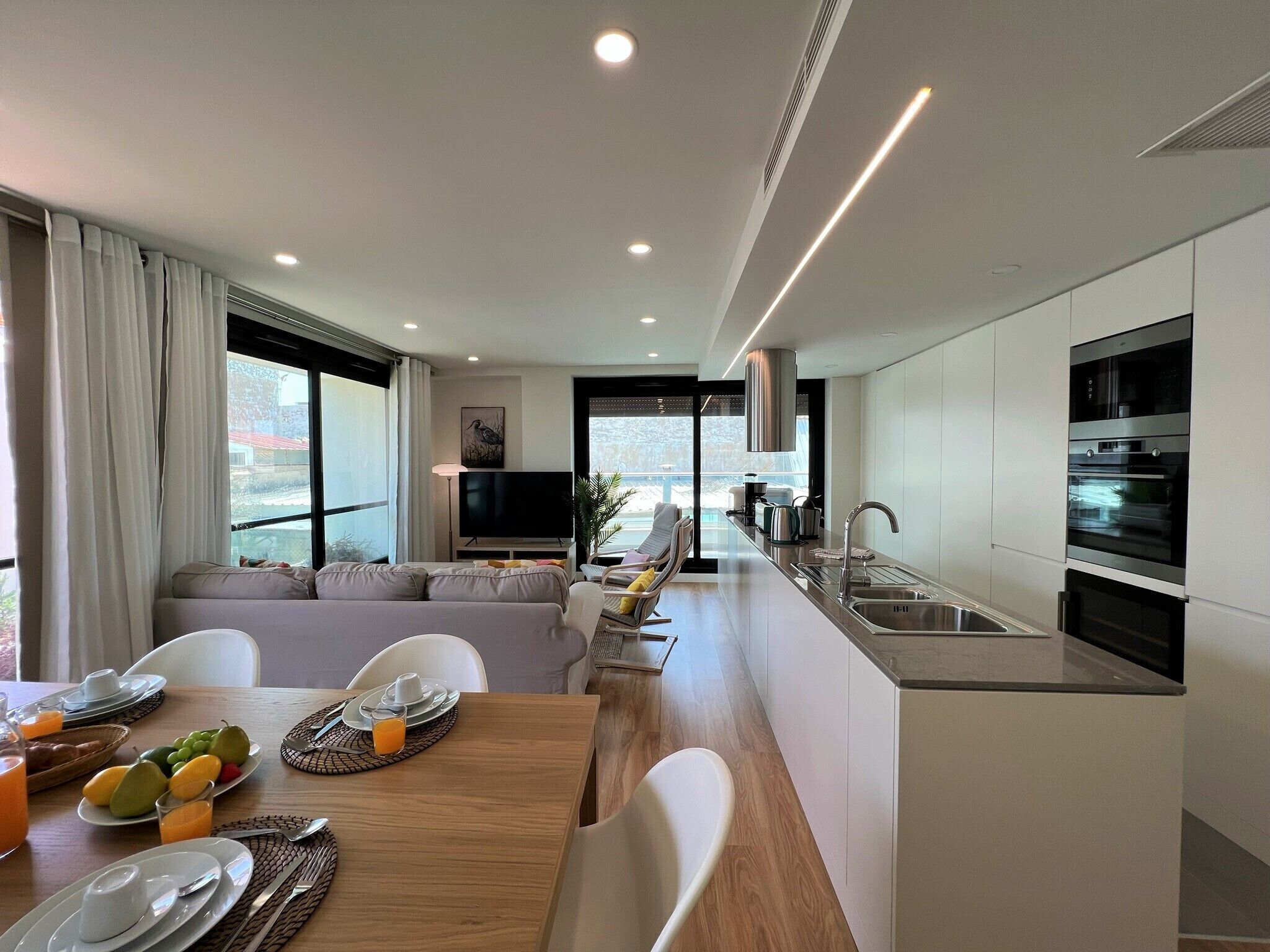 A modern kitchen with built-in appliances, a dining area with a wooden table, and a living area with a sofa and TV.