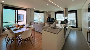 A modern kitchen with a central island, a dining area with a table and chairs, and a living area with a sofa. - FARO DESIGN 1 by HOMING (Faro)