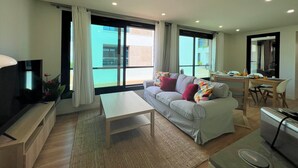 Een moderne woonkamer met een bank, salontafel en televisie. Grote ramen met gordijnen, een eethoek met stoelen en een klein keukentje.
