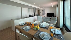 Een moderne eetruimte met een houten tafel gedekt voor het ontbijt, een keuken op de achtergrond en een zithoek met een bank en televisie.
