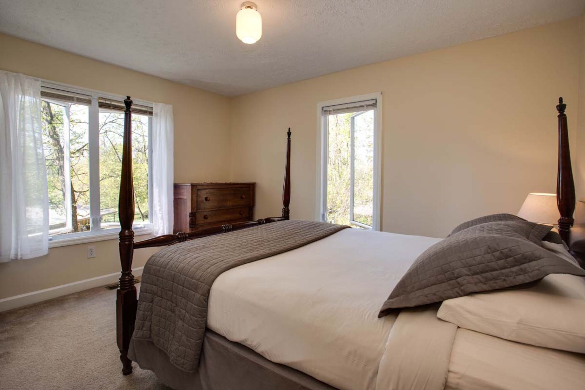 A bedroom with a four-poster bed, a nightstand, and a dresser. There are two windows with curtains.