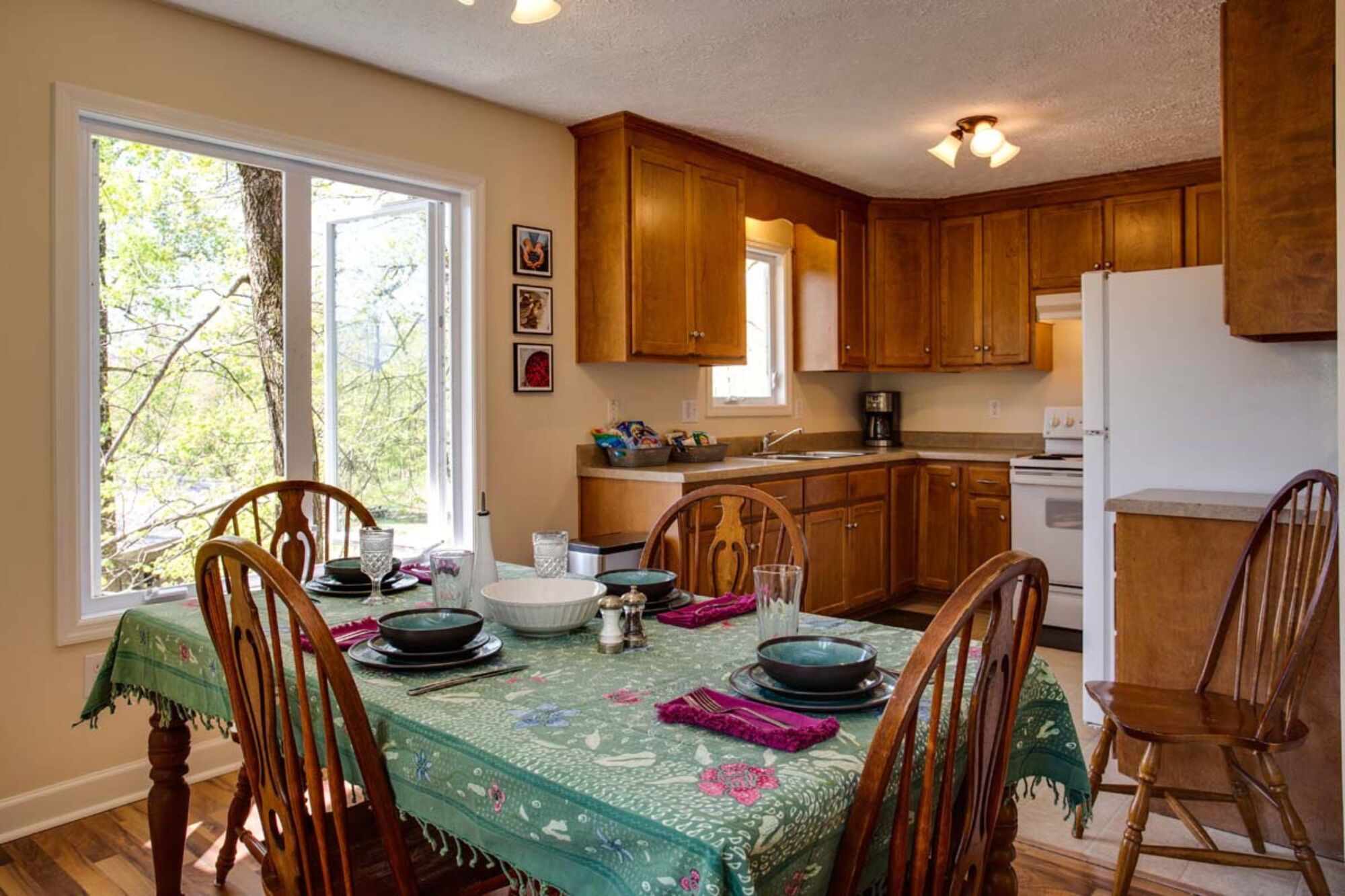 A dining area with a table set for four, wooden cabinets, a refrigerator, and a window with a view of trees.