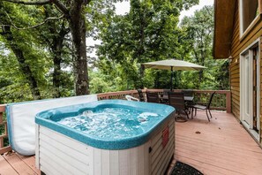 Un bain à remous sur une terrasse en bois, avec une table et des chaises, le tout entouré d’arbres.