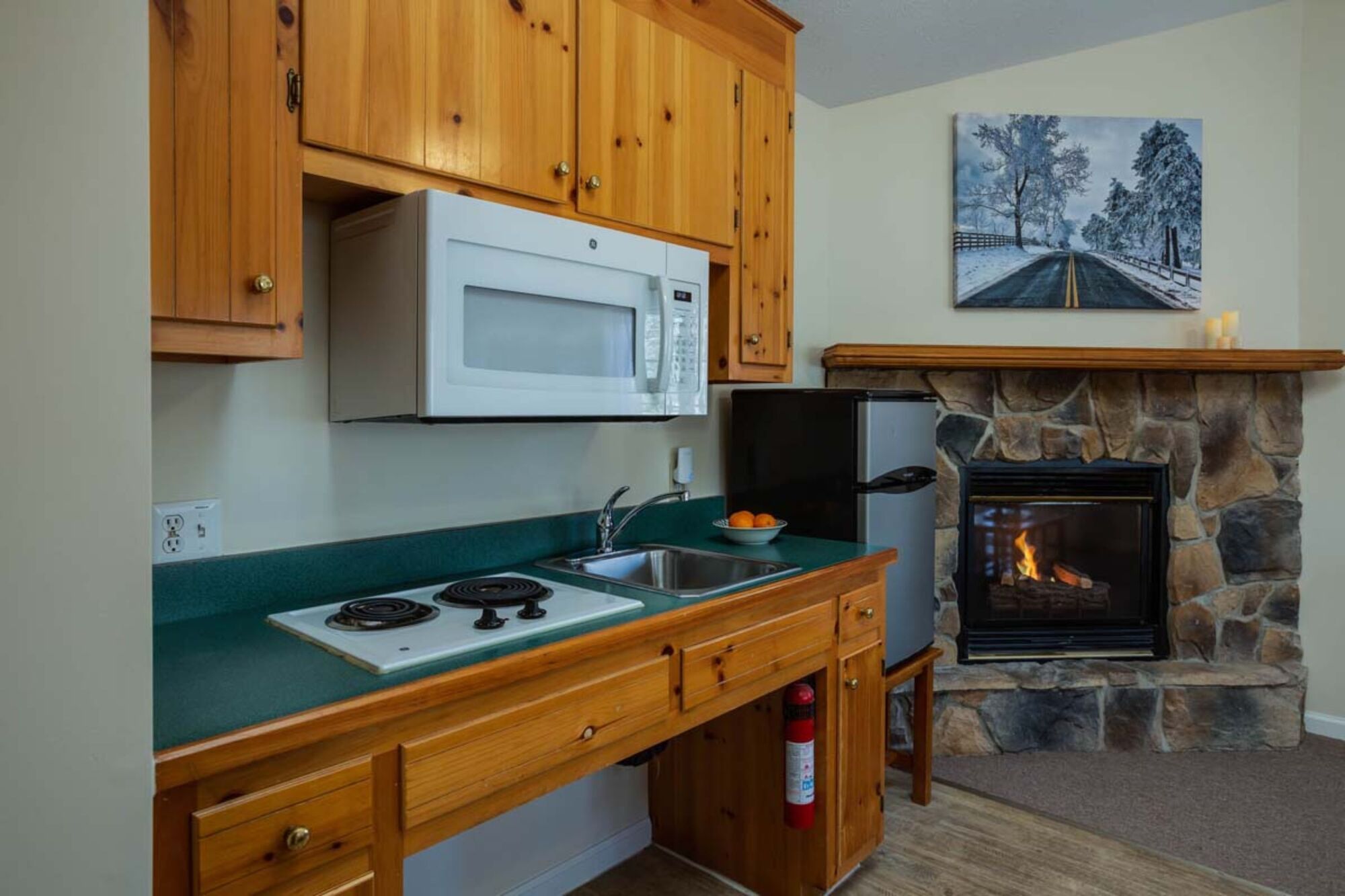 A kitchen with wooden cabinets, a microwave, a stove, a sink, and a stone fireplace with a fire.