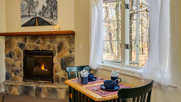 A cozy room with a stone fireplace, a dining table set for two, and a window with a view of trees. - White Rock Cabin--1BR, ADA, kitchenette, fireplace (Lyndhurst)