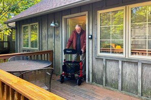 A wooden deck with a table and chairs, a covered patio area, and a house with a door open. - White Rock Cabin--1BR, ADA, kitchenette, fireplace (Lyndhurst)