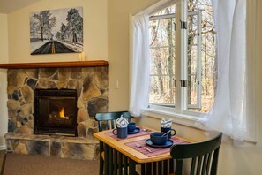 A dining area with a stone fireplace, a table set for two, and a window with a view of trees. - White Rock Cabin--1BR, ADA, kitchenette, fireplace (Lyndhurst)