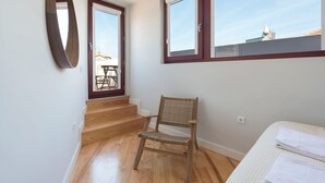 Une pièce avec un plancher en bois, une chaise en osier, un lit avec une literie blanche, un miroir et une fenêtre donnant sur des immeubles.