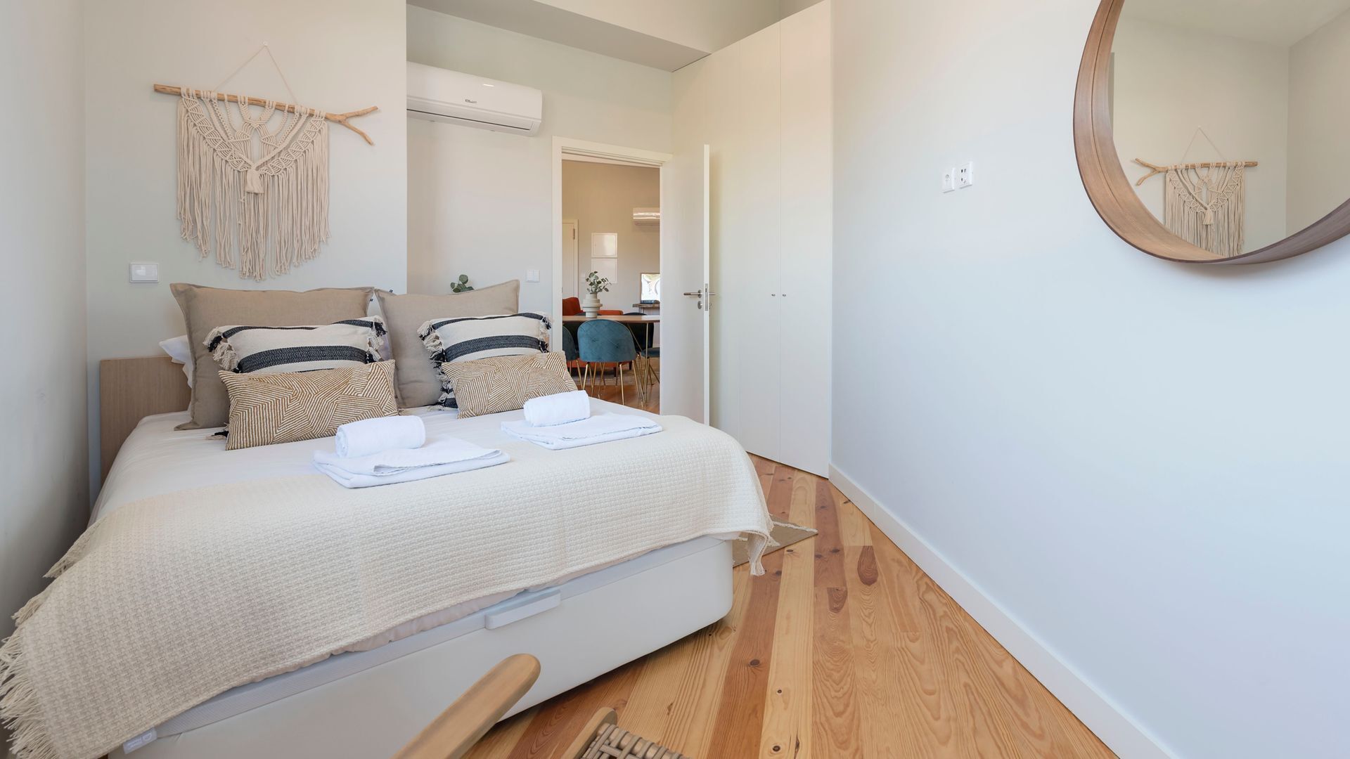 A bedroom with a bed, pillows, a bedside table, a mirror, and a macrame wall hanging.