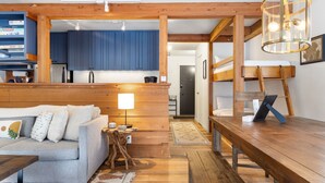 Una sala de estar acogedora con un sofá, una mesa de centro de madera y una mesa de comedor. Se ve una cocina con armarios azules y vigas de madera al fondo.