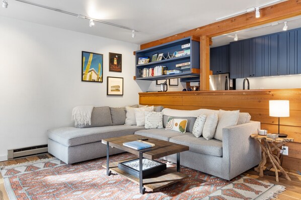 A living room with a grey sofa, a coffee table, a patterned rug, a bookshelf, and a wall-mounted shelf.