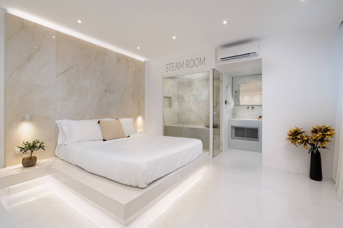 A modern bedroom with a large bed, a glass door leading to a bathroom, and a sign indicating a steam room.