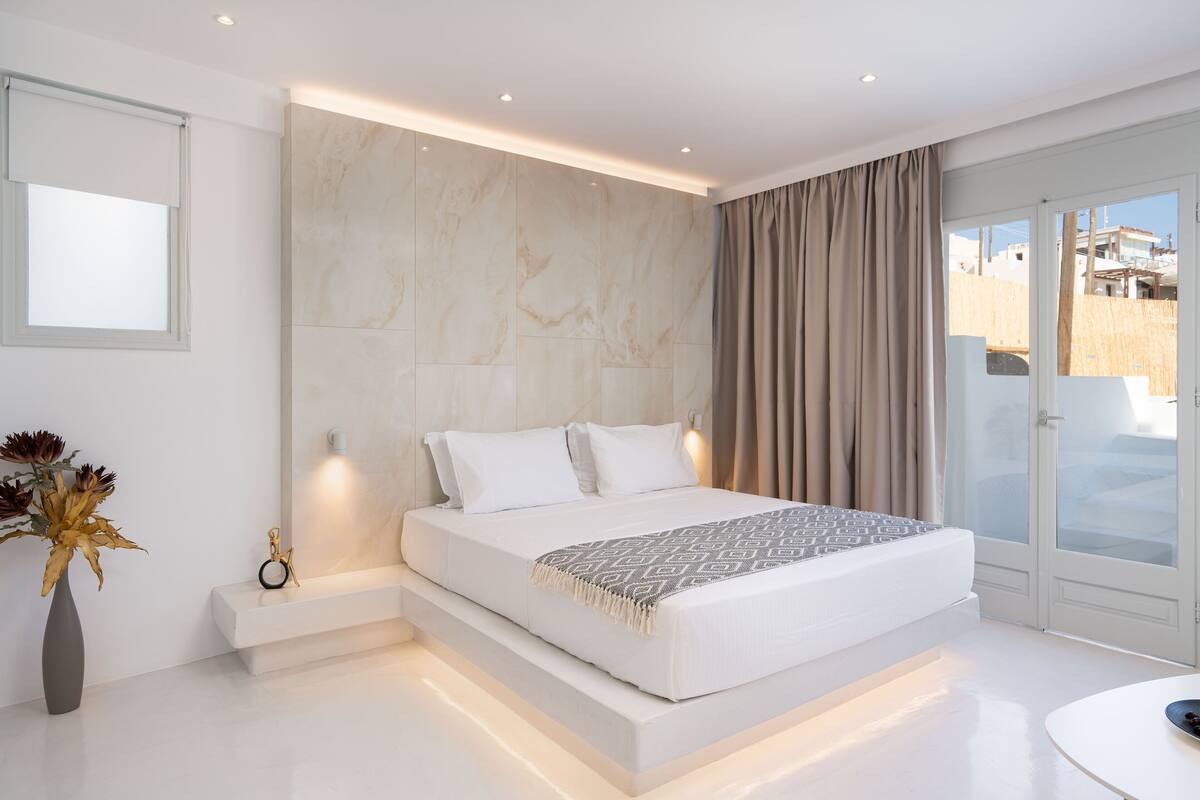 A modern bedroom with a large bed, a bedside table, a vase with flowers, and a view of the outside.