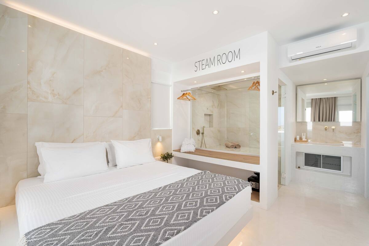 A modern hotel room with a large bed, a steam room, and a bathroom with a mirror and sink.