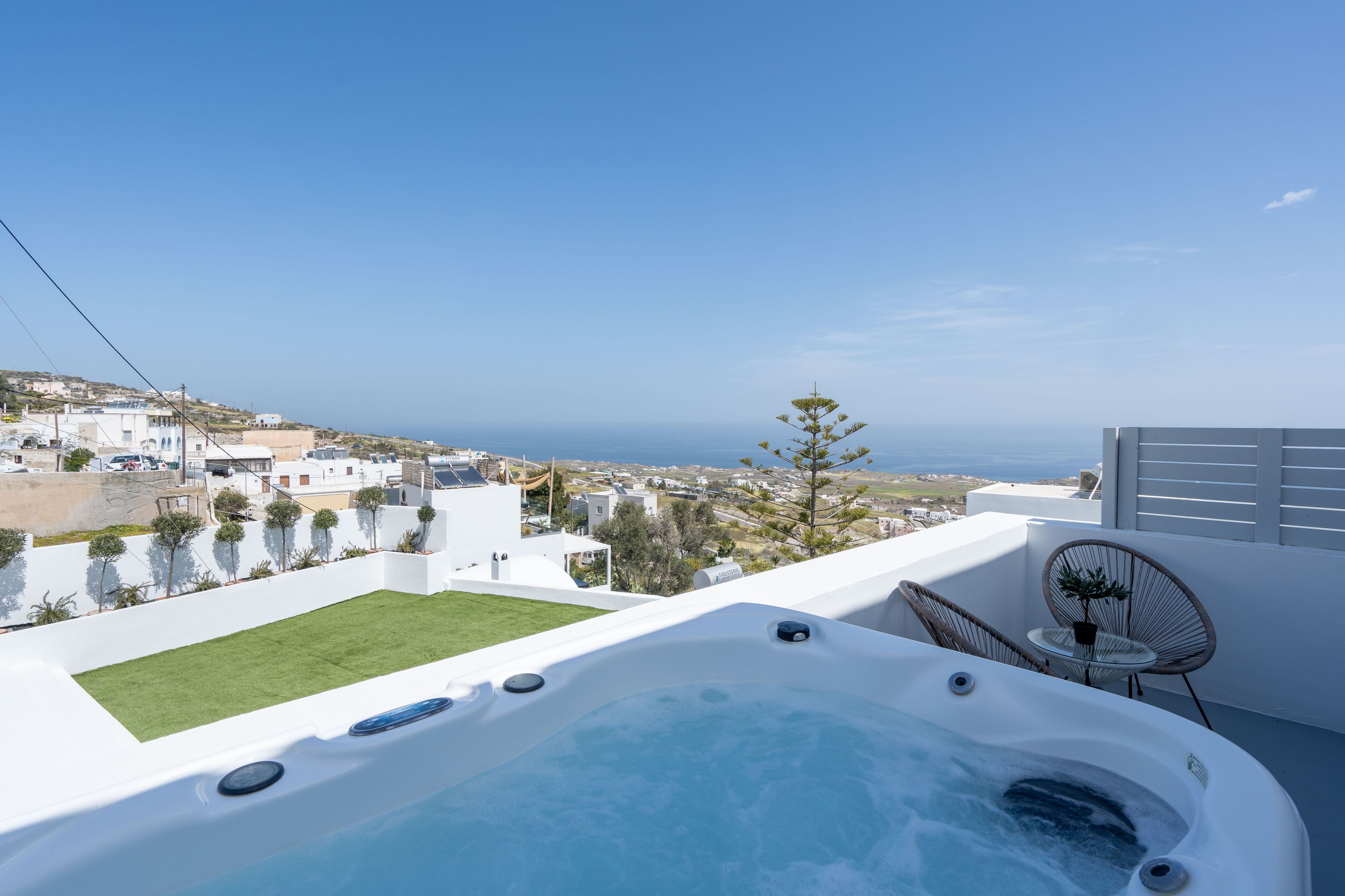 A rooftop terrace with a hot tub, a small table, and two chairs, overlooking a coastal town and the sea.