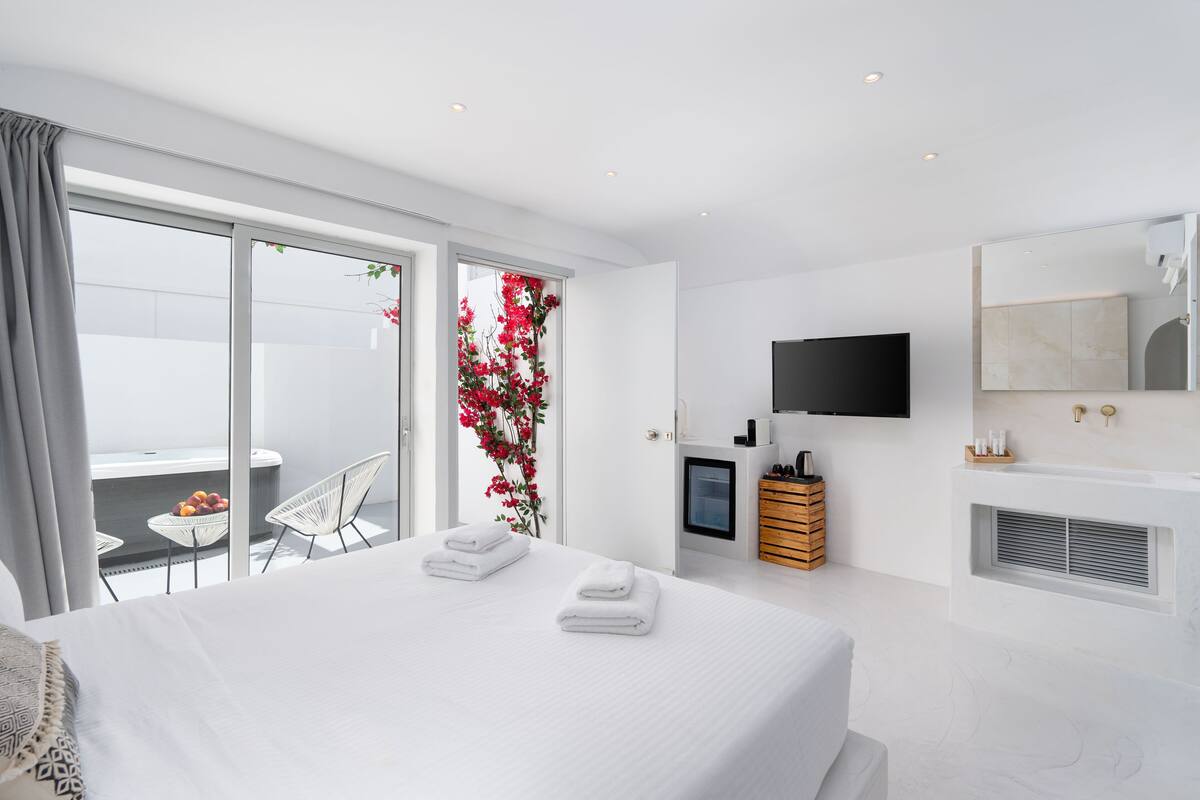 A modern hotel room with a large bed, a flat-screen TV, a bathroom with a bathtub, and a balcony with a table and chairs.