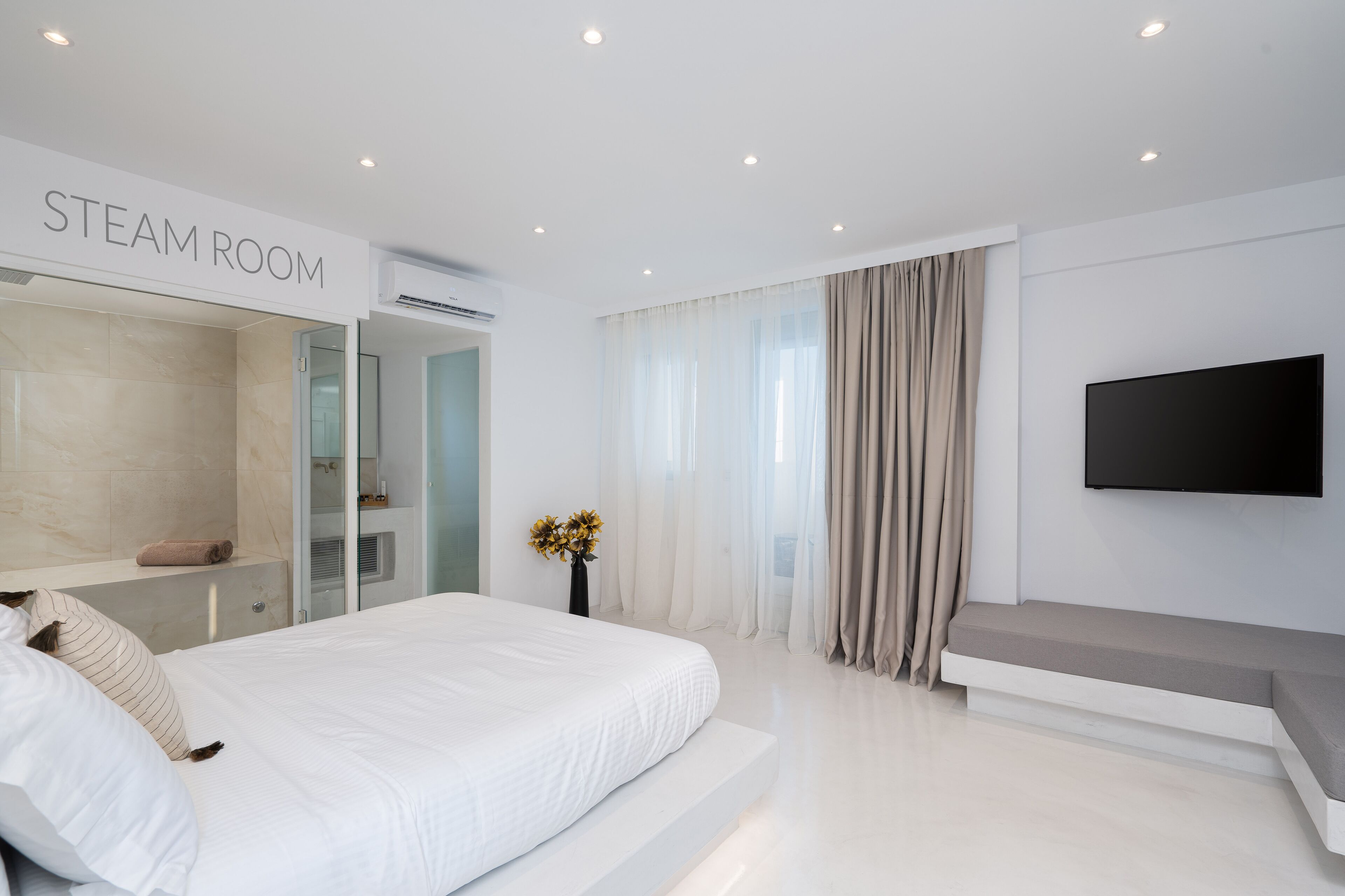 A modern hotel room with a large bed, a TV mounted on the wall, and a mirror with a 'STEAM ROOM' sign.