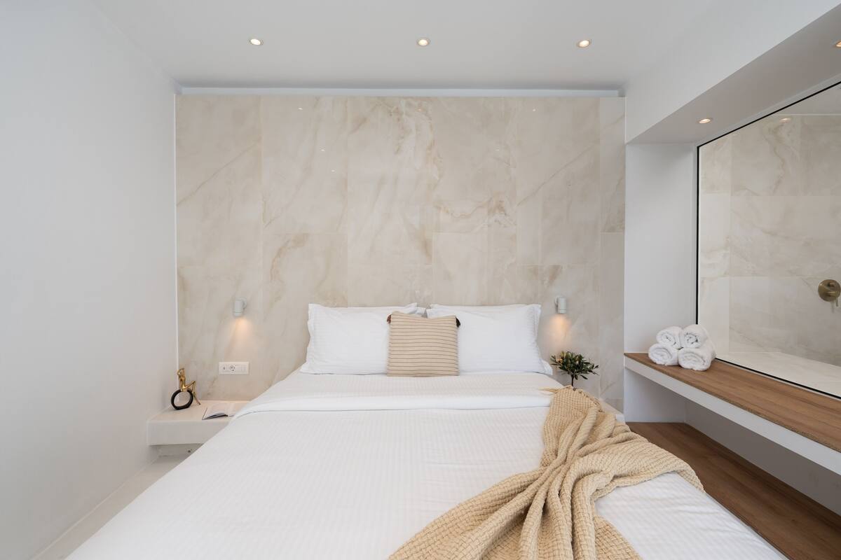 A modern bedroom with a large bed, a bedside table, a nightstand, a mirror, and a wooden shelf.