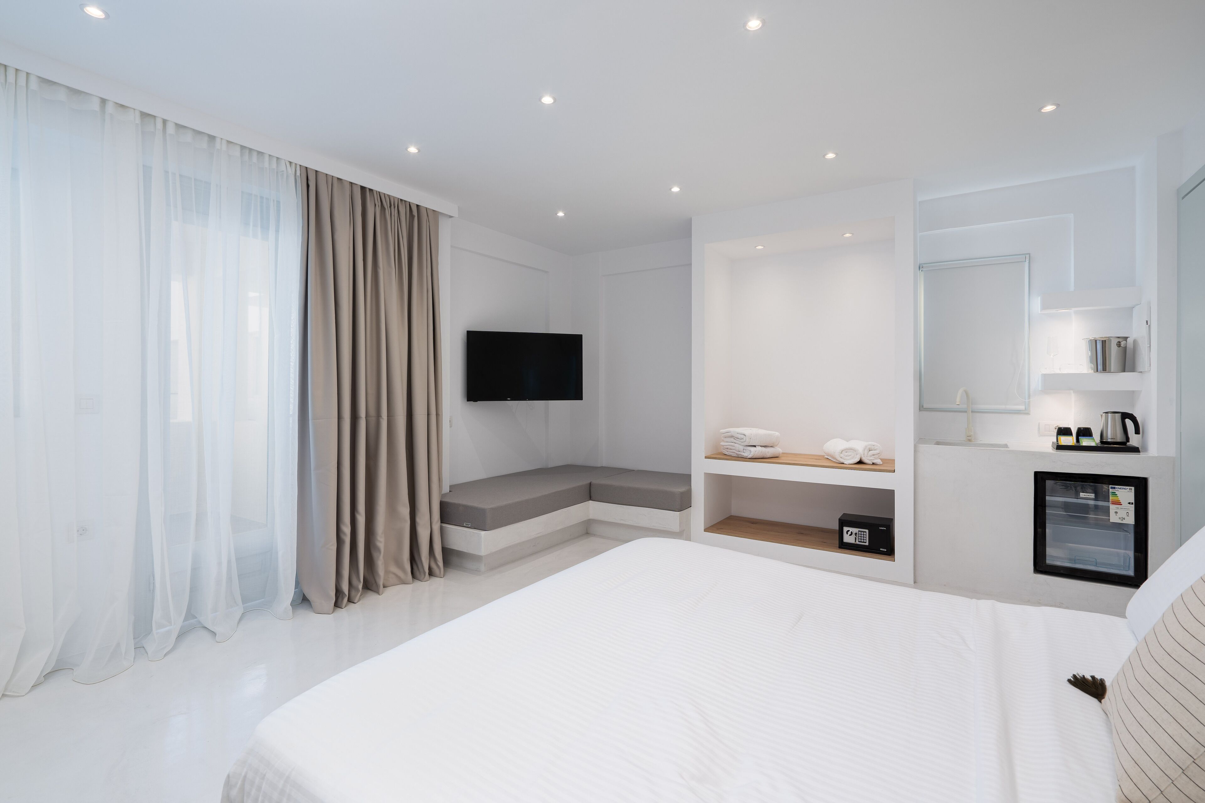 Una camera d'albergo moderna con un letto grande, una TV a schermo piatto montata a parete, una panca integrata con spazio contenitore, un mini-frigorifero e una macchina per il caffè.