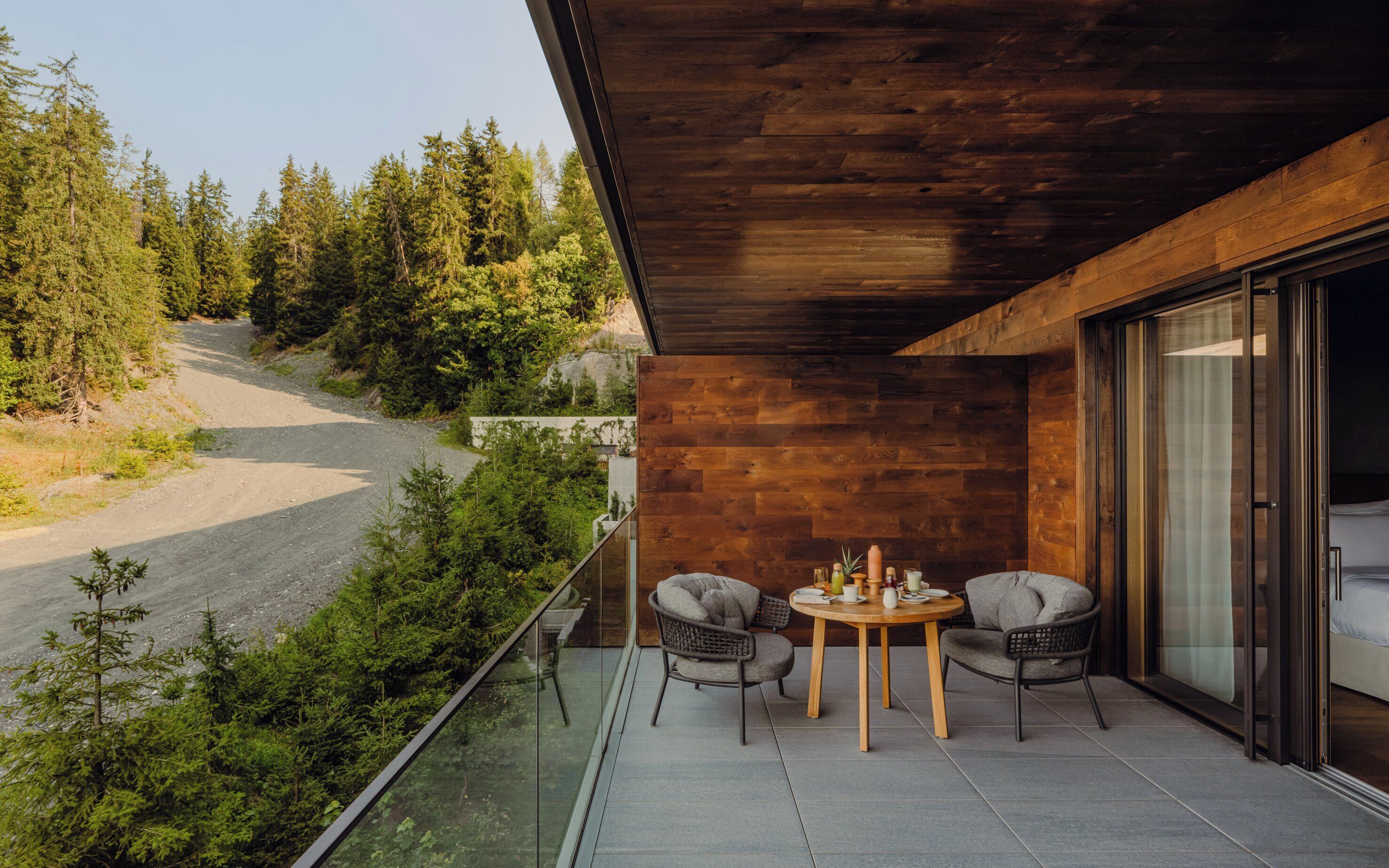 A balcony with a wooden ceiling overlooking a forested area. There is a round table with two chairs and a small potted plant.