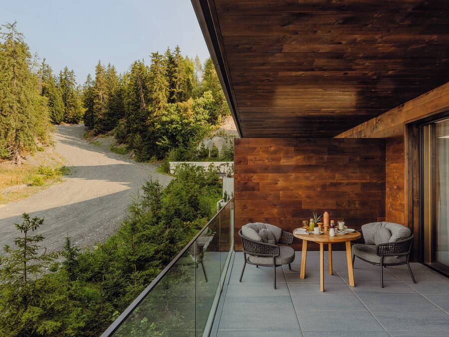 A balcony with a wooden ceiling overlooking a forested area. There is a round table with two chairs and a small potted plant.