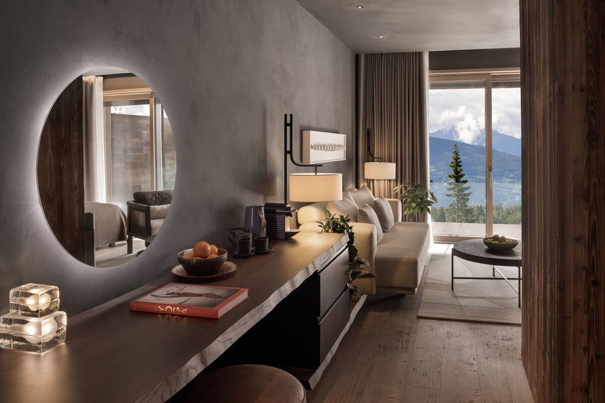 A modern hotel room with a large window offering a mountain view, a cozy seating area with a sofa and armchairs, a wooden dining table with a bowl of fruit, and a small side table with a lamp and decorative items.