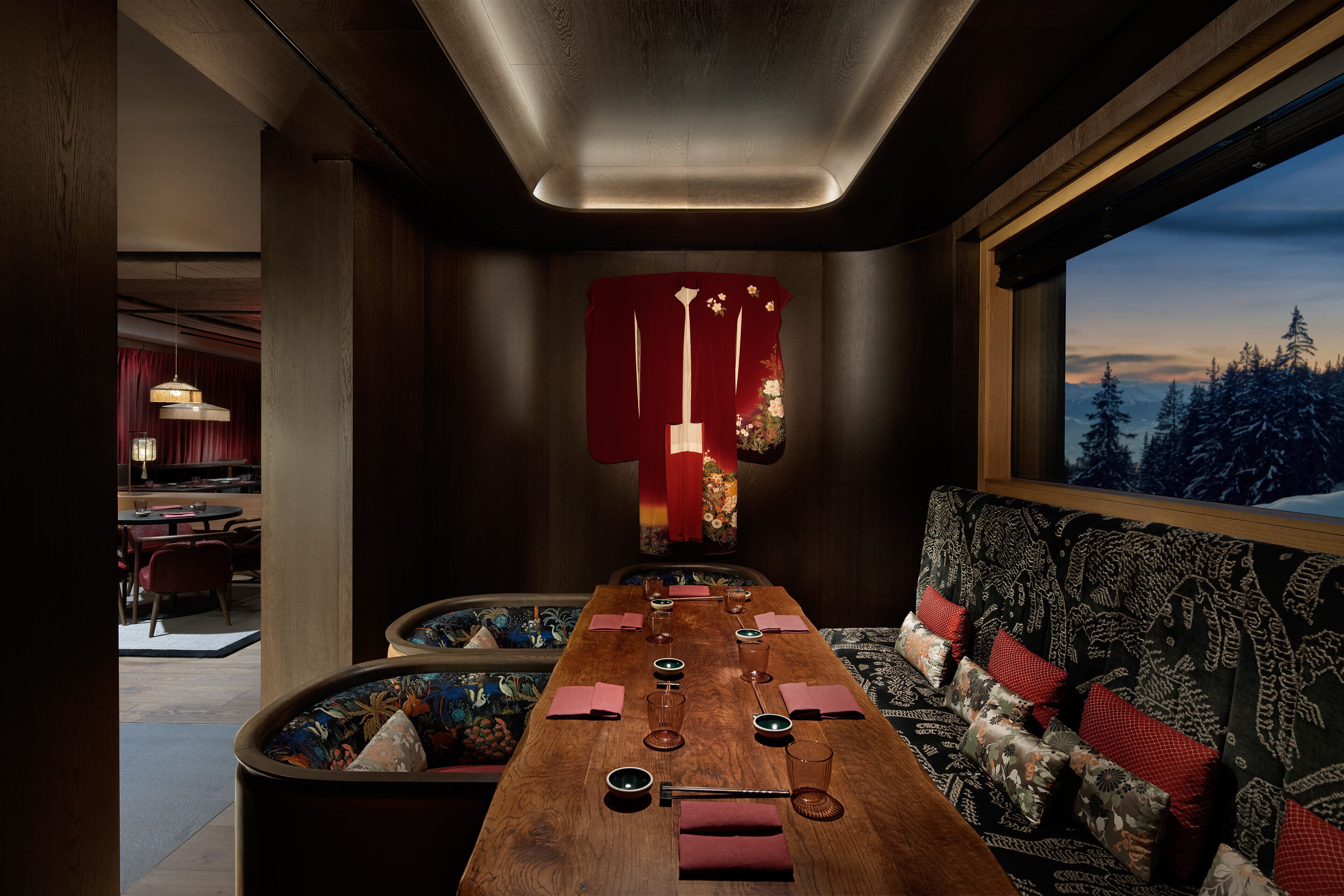 A dining area with a long wooden table set for a meal, a sofa with patterned cushions, and a view of a snowy forest through a window.