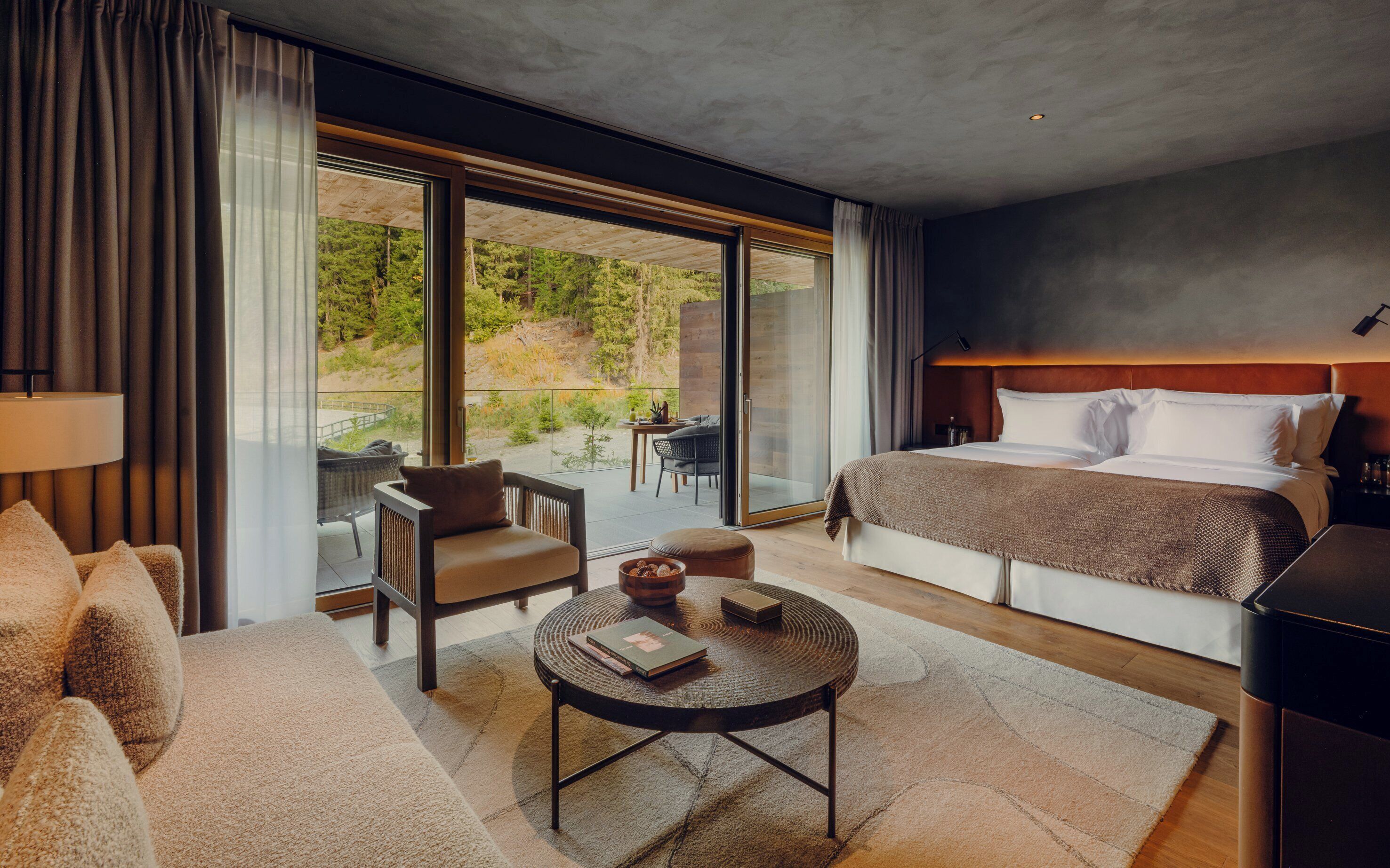 A modern hotel room with a large bed, a sofa, a small round table, and a balcony with a view of greenery.