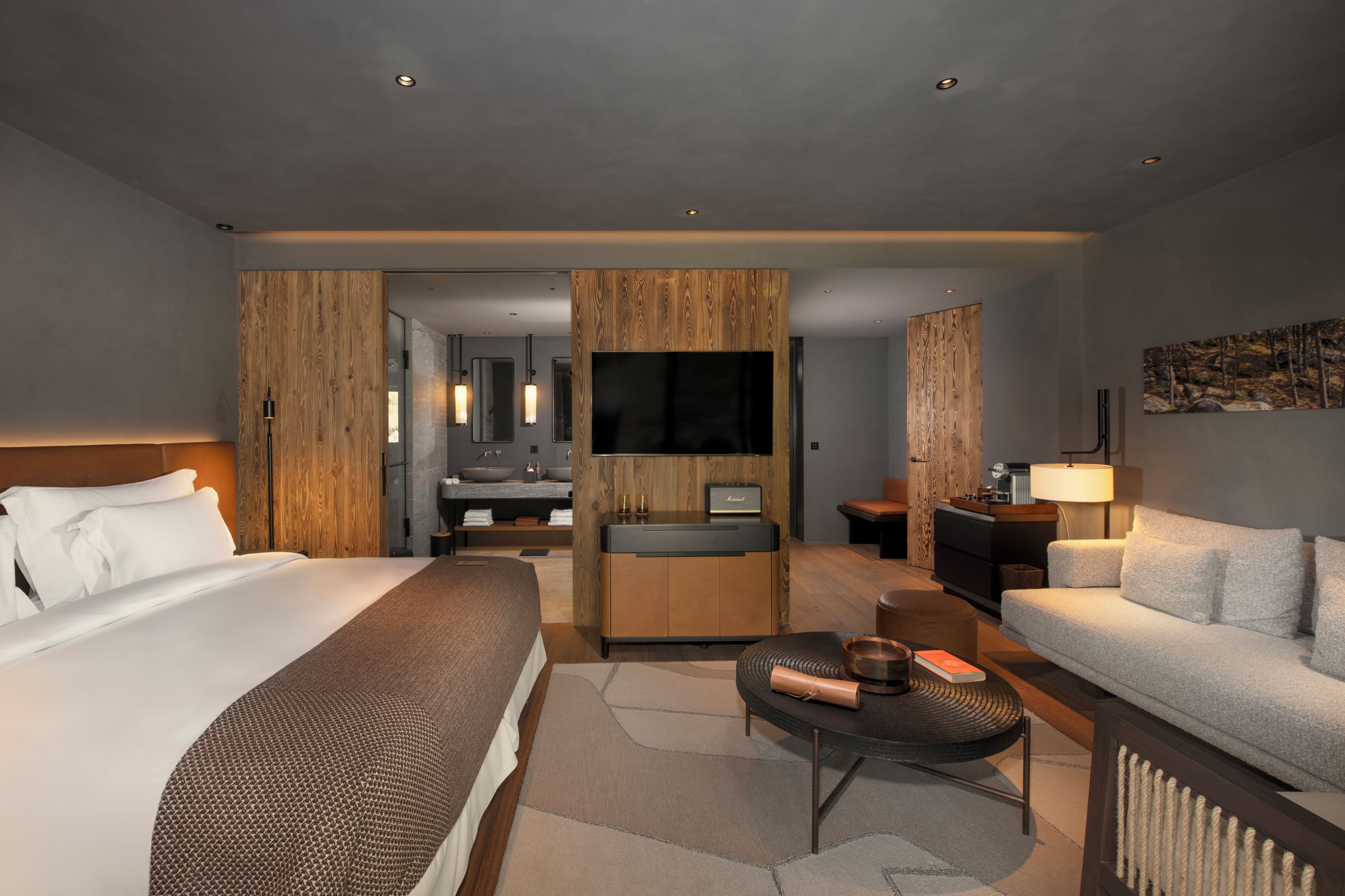 A modern hotel room with a large bed, a sitting area with a sofa and coffee table, a wooden accent wall, a TV, and a bathroom with a sink and mirror.