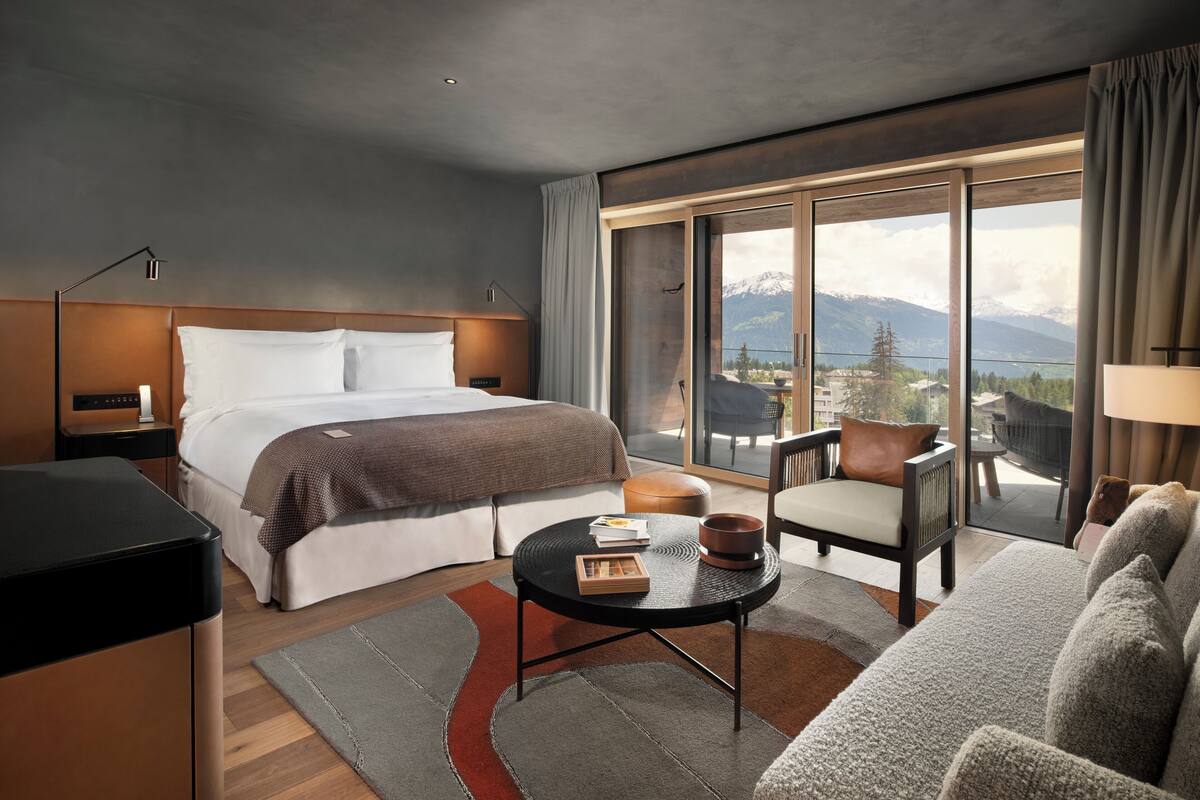 A modern hotel room with a large bed, a round coffee table, a sofa, and a view of mountains through a sliding glass door.