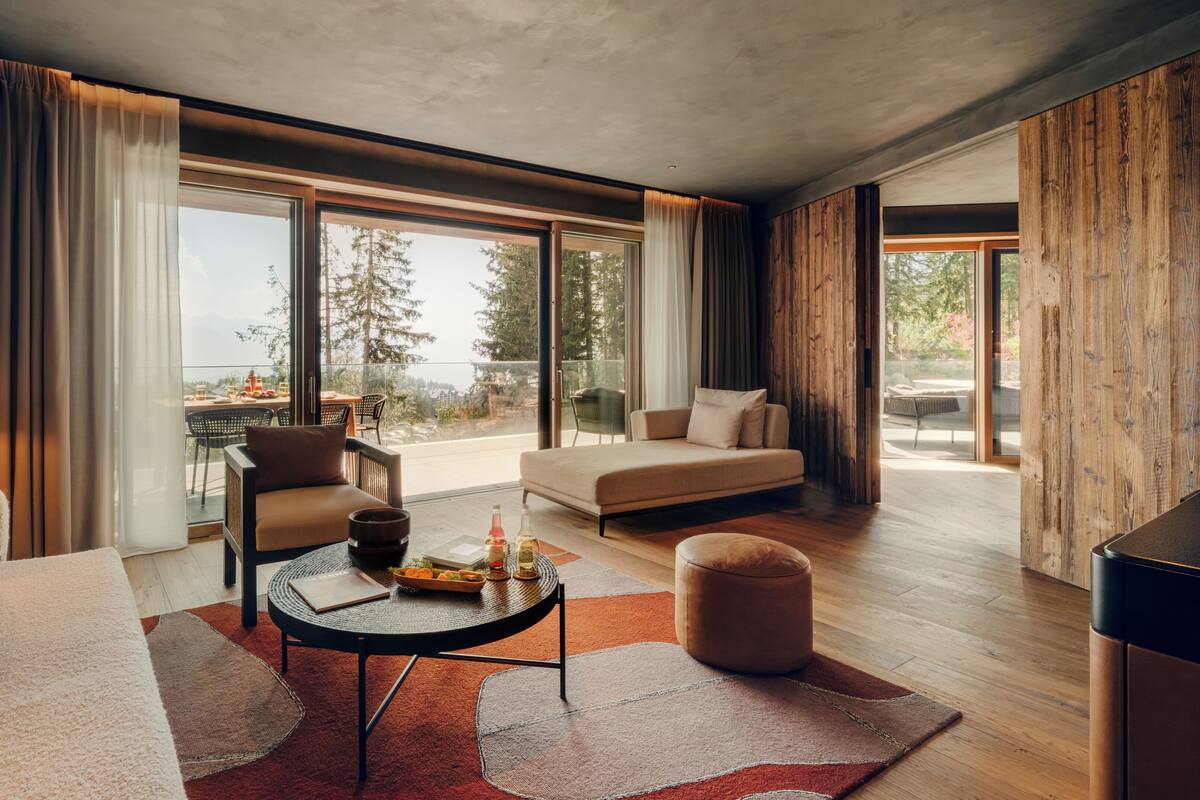 A modern living room with a sofa, armchair, and ottoman, a round coffee table with snacks, and a view of a dining area and trees through large windows.